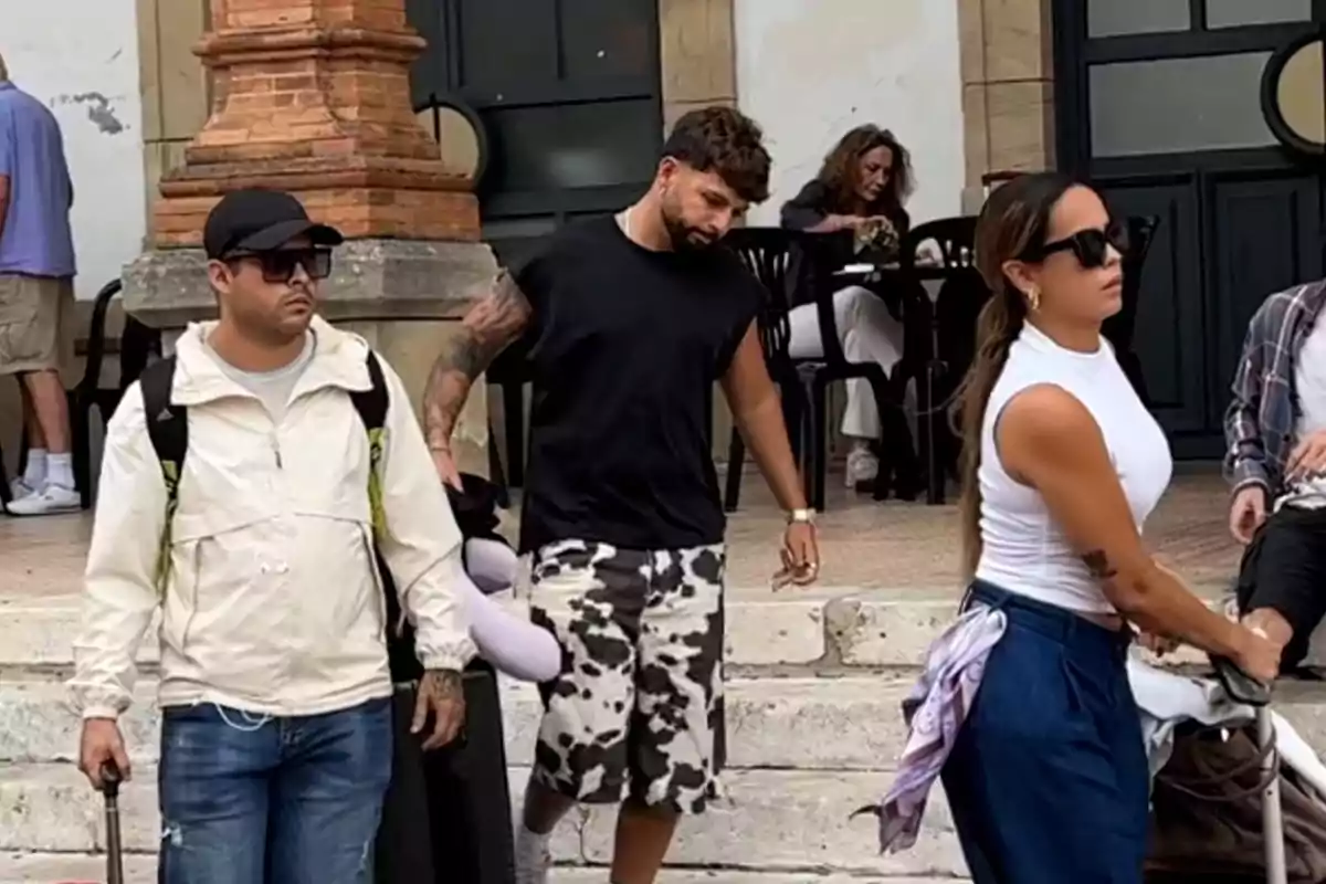 Tres persones caminen pel carrer amb maletes mentre una dona està asseguda al fons en una taula. Tres persones caminen pel carrer amb maletes mentre una dona està asseguda al fons en una taula.