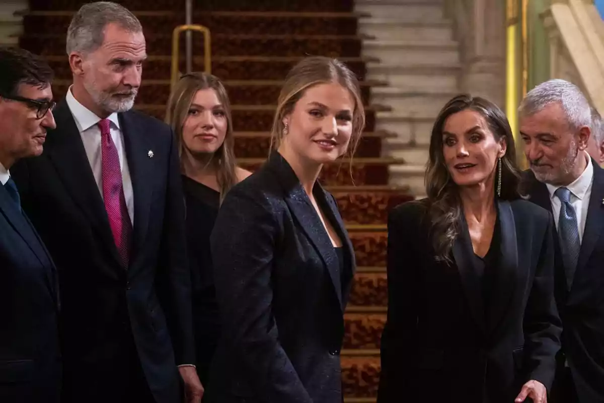 Grupo de personas elegantemente vestidas conversando y sonriendo en el interior de un edificio con una escalera alfombrada al fondo Grupo de personas elegantemente vestidas conversando y sonriendo en el interior de un edificio con una escalera alfombrada al fondo