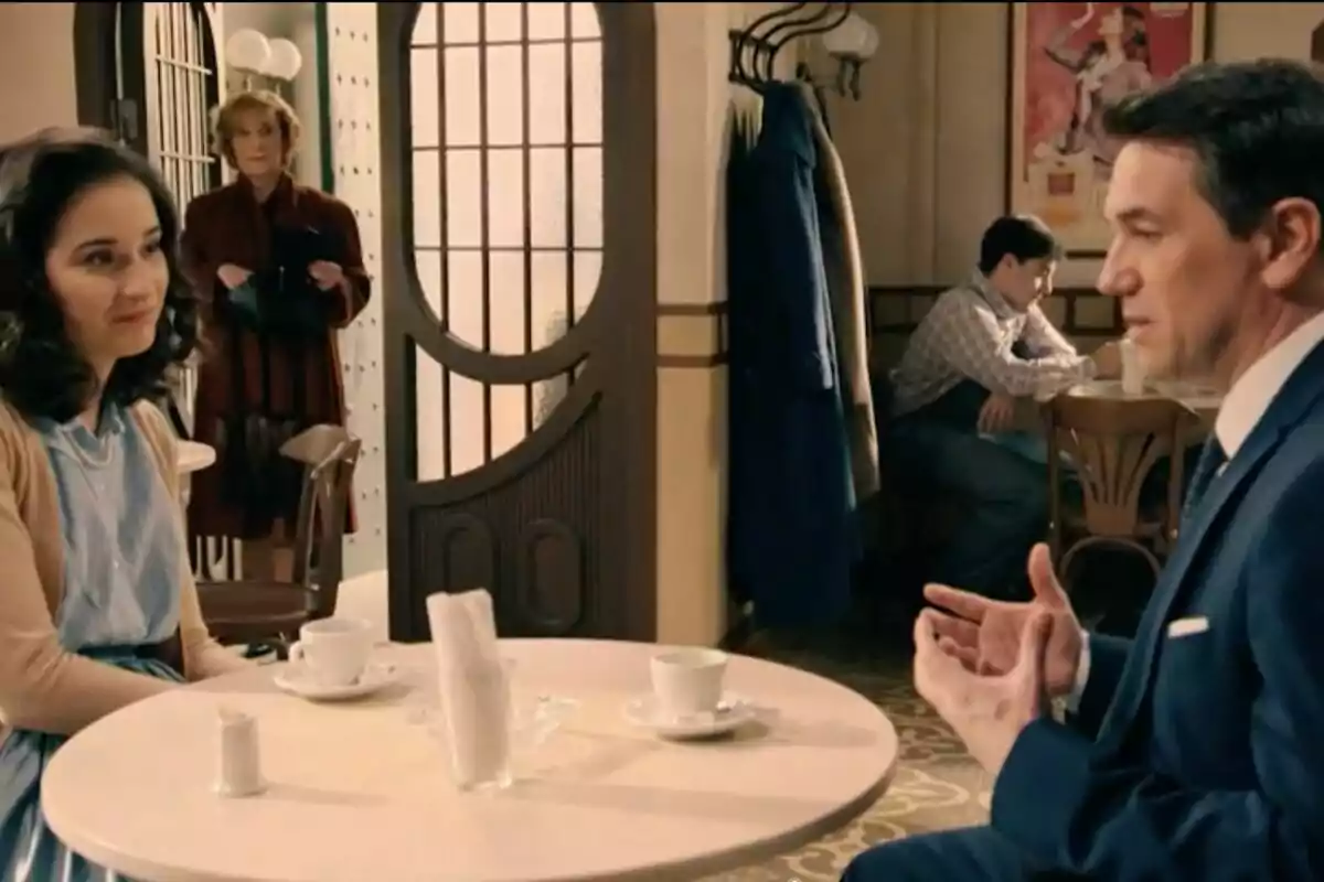 Dues persones, que són Gabriel i Cristina, conversen en una taula de cafeteria mentre una dona observa des del fons i un home està assegut sol en una altra taula. Dues persones, que són Gabriel i Cristina, conversen en una taula de cafeteria mentre una dona observa des del fons i un home està assegut sol en una altra taula.