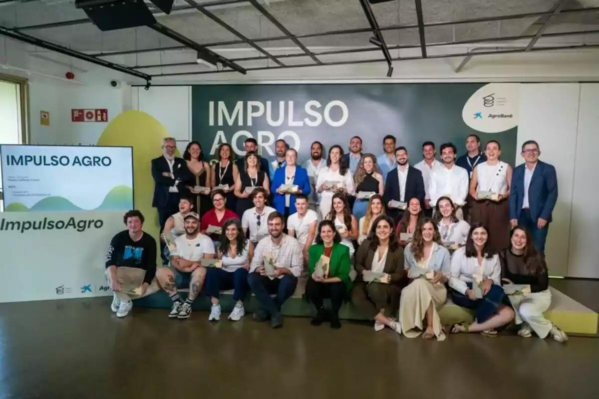 Un grup nombrós de persones somriu posant per a una foto de grup en un esdeveniment anomenat Impuls Agro, amb un fons i una pantalla que mostren el nom de l’esdeveniment. Un grup nombrós de persones somriu posant per a una foto de grup en un esdeveniment anomenat Impuls Agro, amb un fons i una pantalla que mostren el nom de l’esdeveniment.