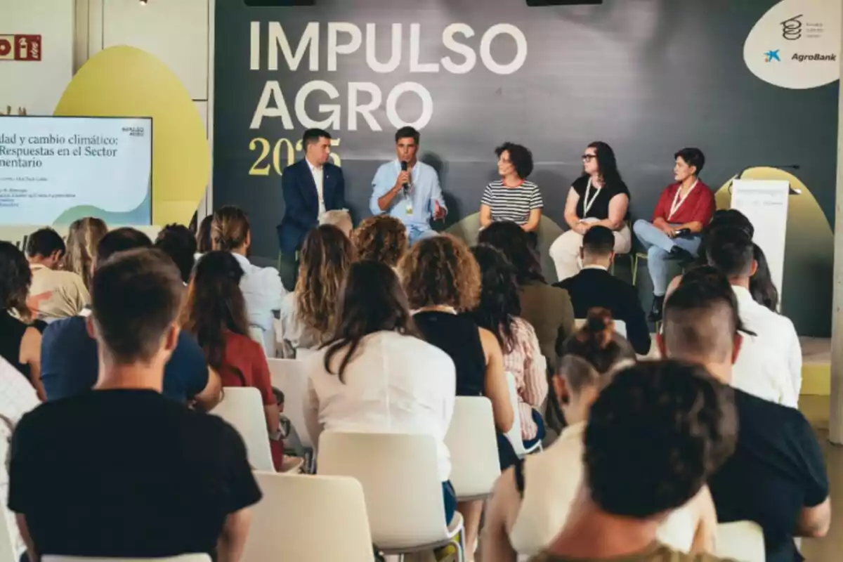 Un grup de persones assisteix a una conferència on cinc ponents estan asseguts a l’escenari participant en una xerrada sobre sostenibilitat i canvi climàtic en el sector alimentari, amb un fons que diu Impuls Agro 2023. Un grup de persones assisteix a una conferència on cinc ponents estan asseguts a l’escenari participant en una xerrada sobre sostenibilitat i canvi climàtic en el sector alimentari, amb un fons que diu Impuls Agro 2023.