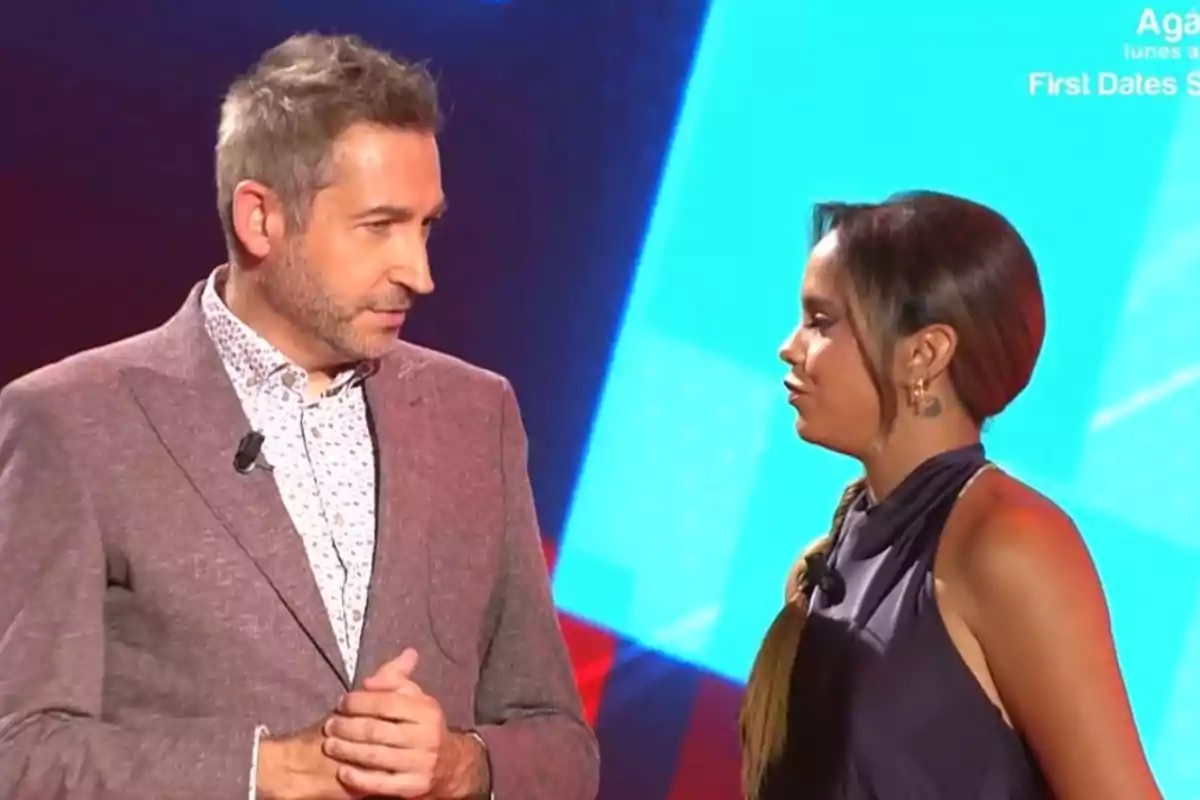 Un home amb jaqueta clara i una dona amb vestit fosc conversen en un plató de televisió amb fons blau. Un home amb jaqueta clara i una dona amb vestit fosc conversen en un plató de televisió amb fons blau.