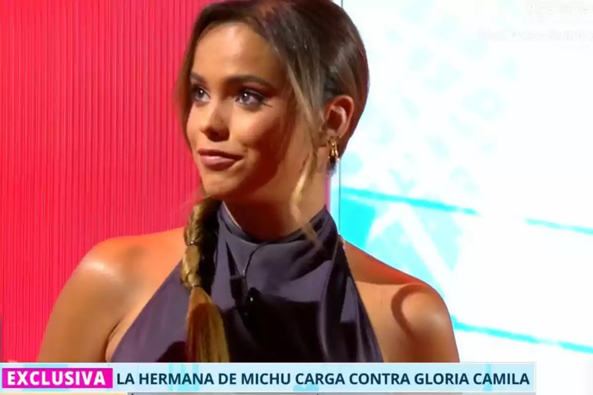 Dona jove amb cua i vestit gris en un plató de televisió amb fons vermell i blau, apareix un rètol informatiu a la part inferior Dona jove amb cua i vestit gris en un plató de televisió amb fons vermell i blau, apareix un rètol informatiu a la part inferior