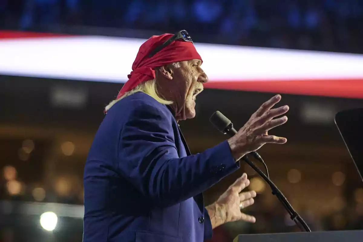 Long-haired man with a mustache wearing a red bandana and a blue jacket, speaking passionately into a microphone on a stage Long-haired man with a mustache wearing a red bandana and a blue jacket, speaking passionately into a microphone on a stage