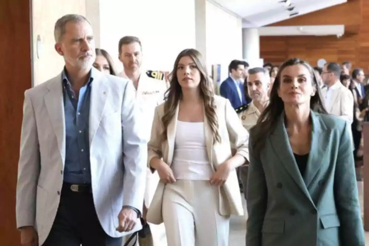 Tres persones vestides de manera formal caminen per un passadís acompanyades d’altres persones al fons. Tres persones vestides de manera formal caminen per un passadís acompanyades d’altres persones al fons.