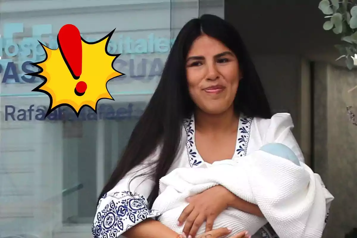 Dona de cabells llargs i foscos somriu mentre sosté un nadó embolicat amb una manta blanca, amb un gràfic d’exclamació groc i vermell sobre el costat esquerre de la imatge.
