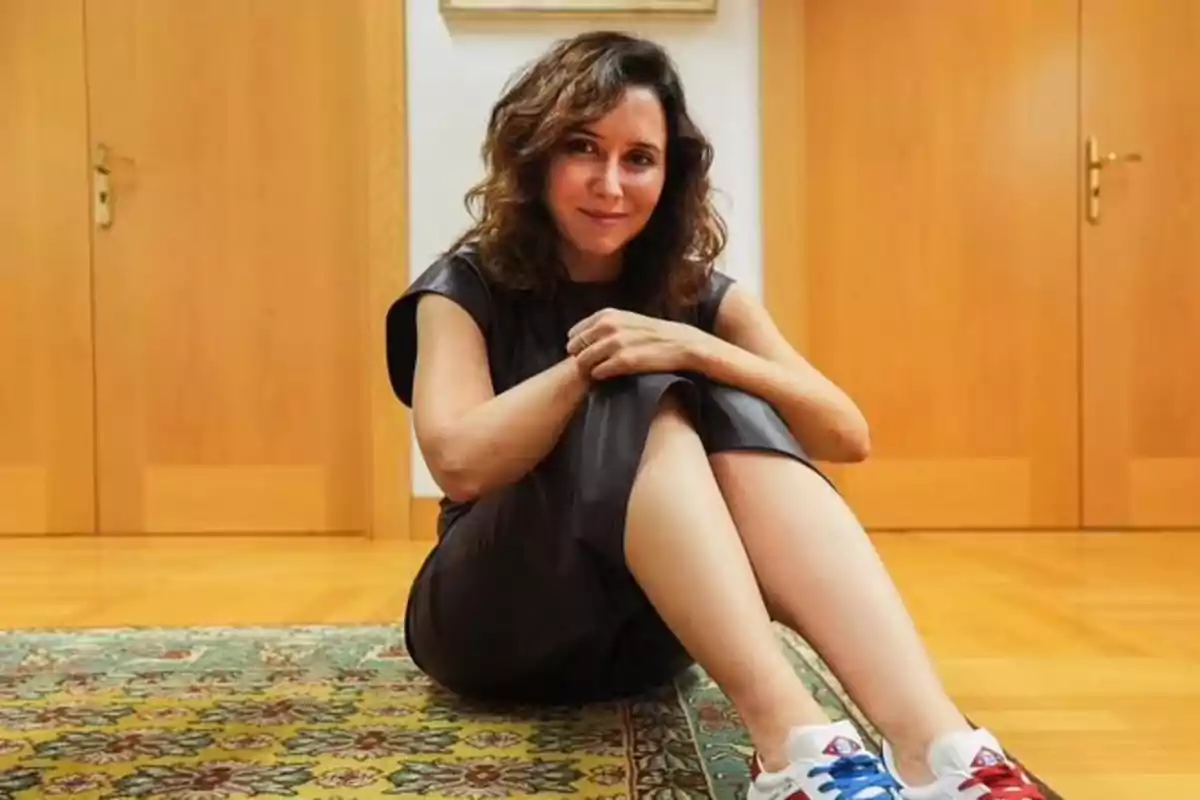 Isabel Díaz Ayuso sentada en el suelo sobre una alfombra colorida con las piernas cruzadas, lleva un vestido oscuro y zapatillas deportivas, al fondo hay puertas de madera. Isabel Díaz Ayuso sentada en el suelo sobre una alfombra colorida con las piernas cruzadas, lleva un vestido oscuro y zapatillas deportivas, al fondo hay puertas de madera.