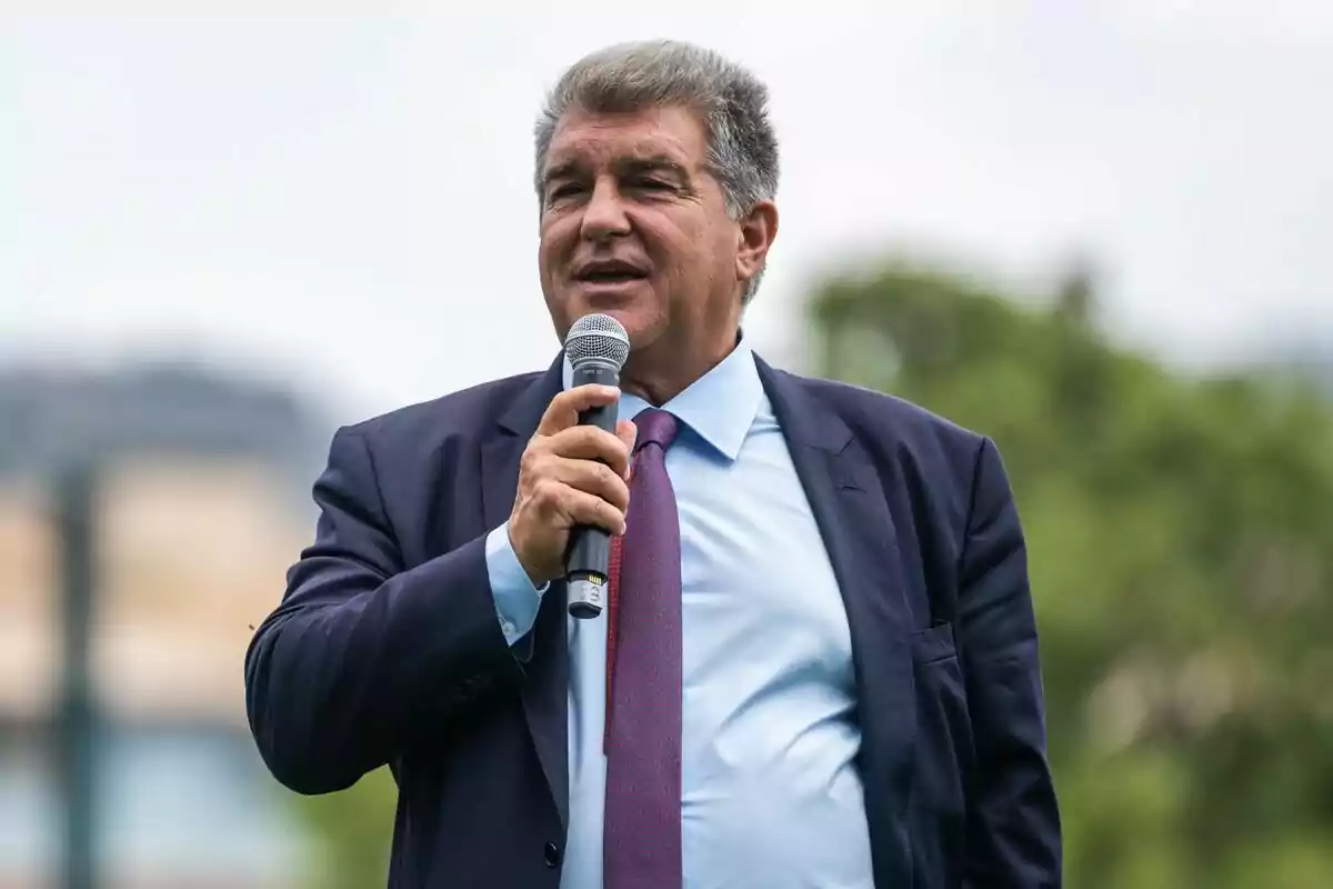 Hombre de cabello canoso con traje azul y corbata roja hablando al aire libre mientras sostiene un micrófono Hombre de cabello canoso con traje azul y corbata roja hablando al aire libre mientras sostiene un micrófono