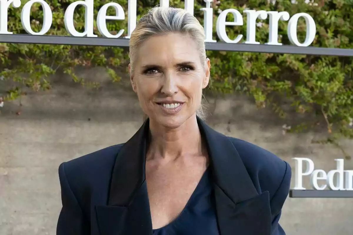 Mujer rubia con el cabello recogido y vestida con un saco azul oscuro sonríe frente a un fondo con letras blancas y vegetación Mujer rubia con el cabello recogido y vestida con un saco azul oscuro sonríe frente a un fondo con letras blancas y vegetación