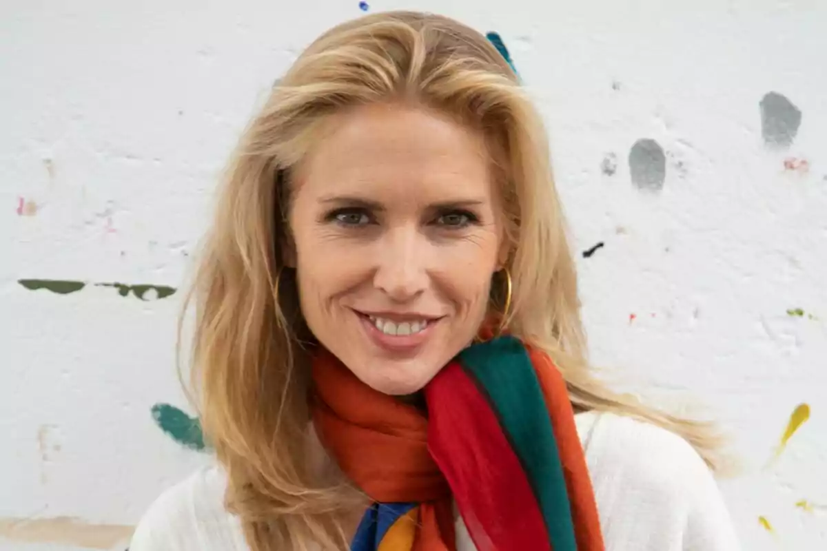 Mujer rubia sonriendo con bufanda colorida frente a una pared blanca con manchas de pintura Mujer rubia sonriendo con bufanda colorida frente a una pared blanca con manchas de pintura