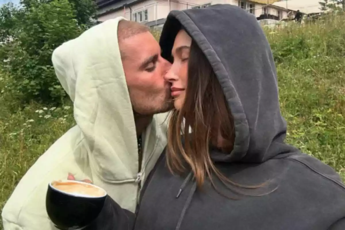 Pareja, formada por Justin Bieber y Hailey Bieber, con sudaderas abrazándose mientras uno de ellos besa la mejilla del otro y sostiene una taza de café en un entorno al aire libre con césped y árboles. Pareja, formada por Justin Bieber y Hailey Bieber, con sudaderas abrazándose mientras uno de ellos besa la mejilla del otro y sostiene una taza de café en un entorno al aire libre con césped y árboles.