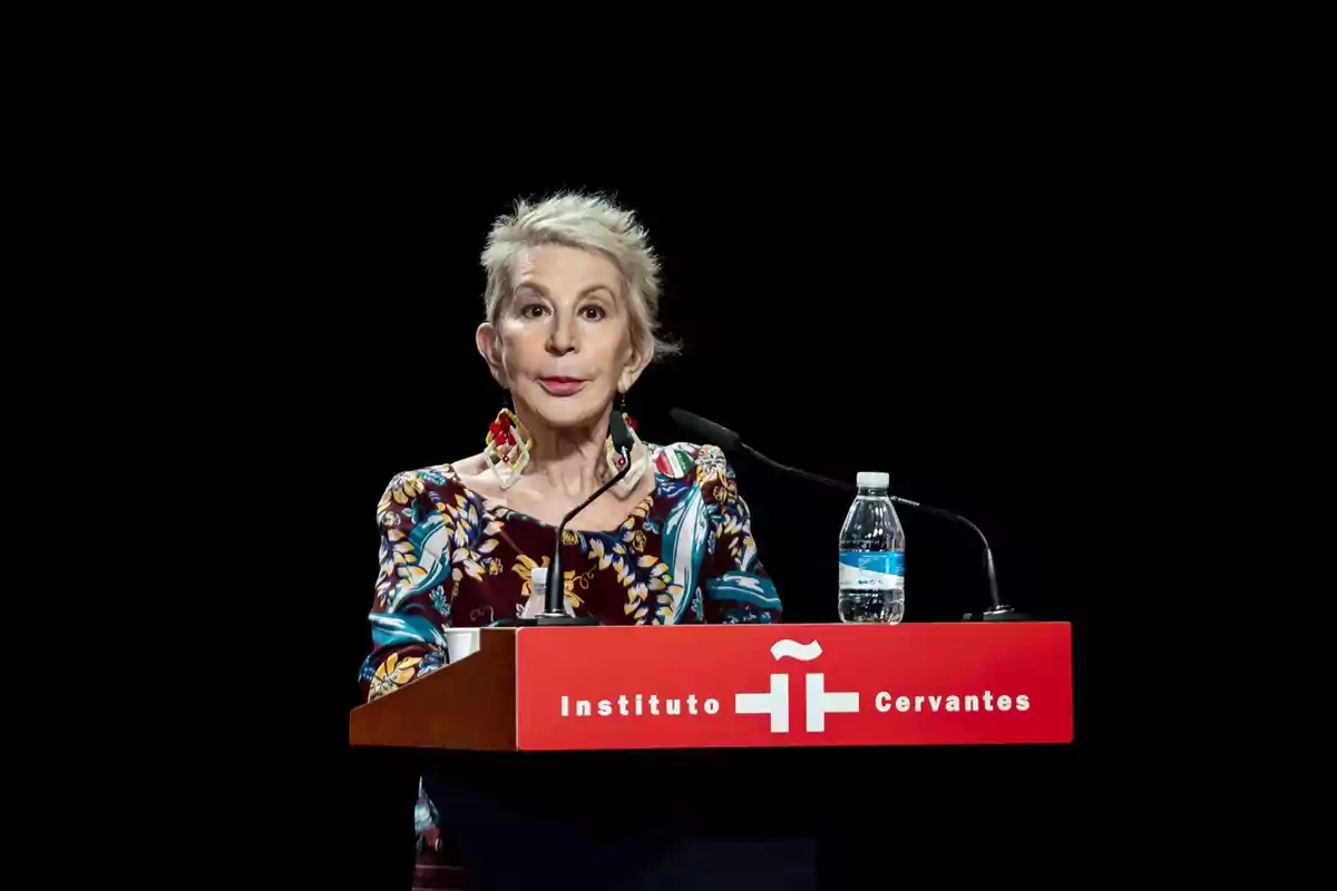 Dona de cabell curt i canós parlant en un faristol de l'Institut Cervantes amb fons negre i una ampolla d'aigua sobre la taula Dona de cabell curt i canós parlant en un faristol de l'Institut Cervantes amb fons negre i una ampolla d'aigua sobre la taula