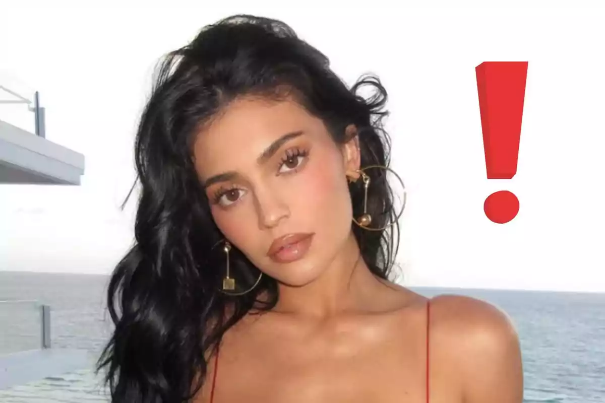 Kylie Jenner de cabello oscuro con pendientes grandes posando frente al mar y un signo de exclamación rojo a su lado