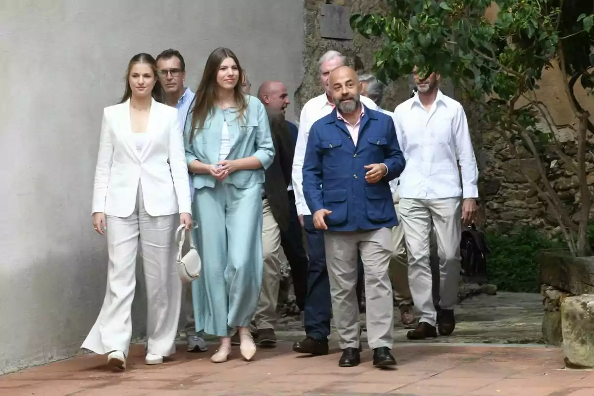 Un grup de persones elegantment vestides camina per un camí de pedra al costat d’una paret i vegetació.