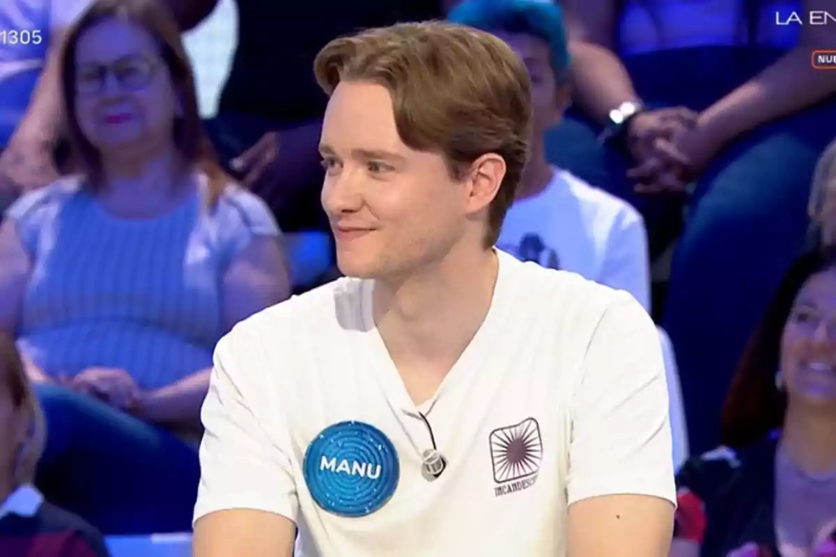 Jove amb samarreta blanca i placa blava que diu Manu participant en un programa de televisió amb públic de fons Jove amb samarreta blanca i placa blava que diu Manu participant en un programa de televisió amb públic de fons