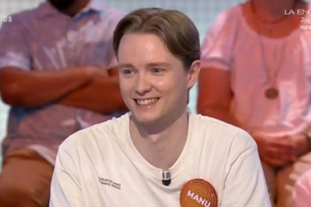 Manu sonriente con camiseta blanca y un gafete que dice Manu participando en un programa de televisión con personas sentadas al fondo Manu sonriente con camiseta blanca y un gafete que dice Manu participando en un programa de televisión con personas sentadas al fondo