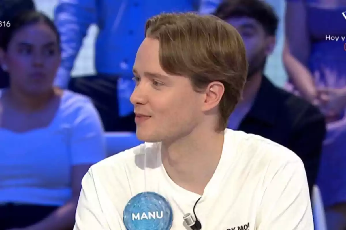 Manu con camiseta blanca y gafete azul que dice Manu participando en un programa de televisión con público al fondo Manu con camiseta blanca y gafete azul que dice Manu participando en un programa de televisión con público al fondo