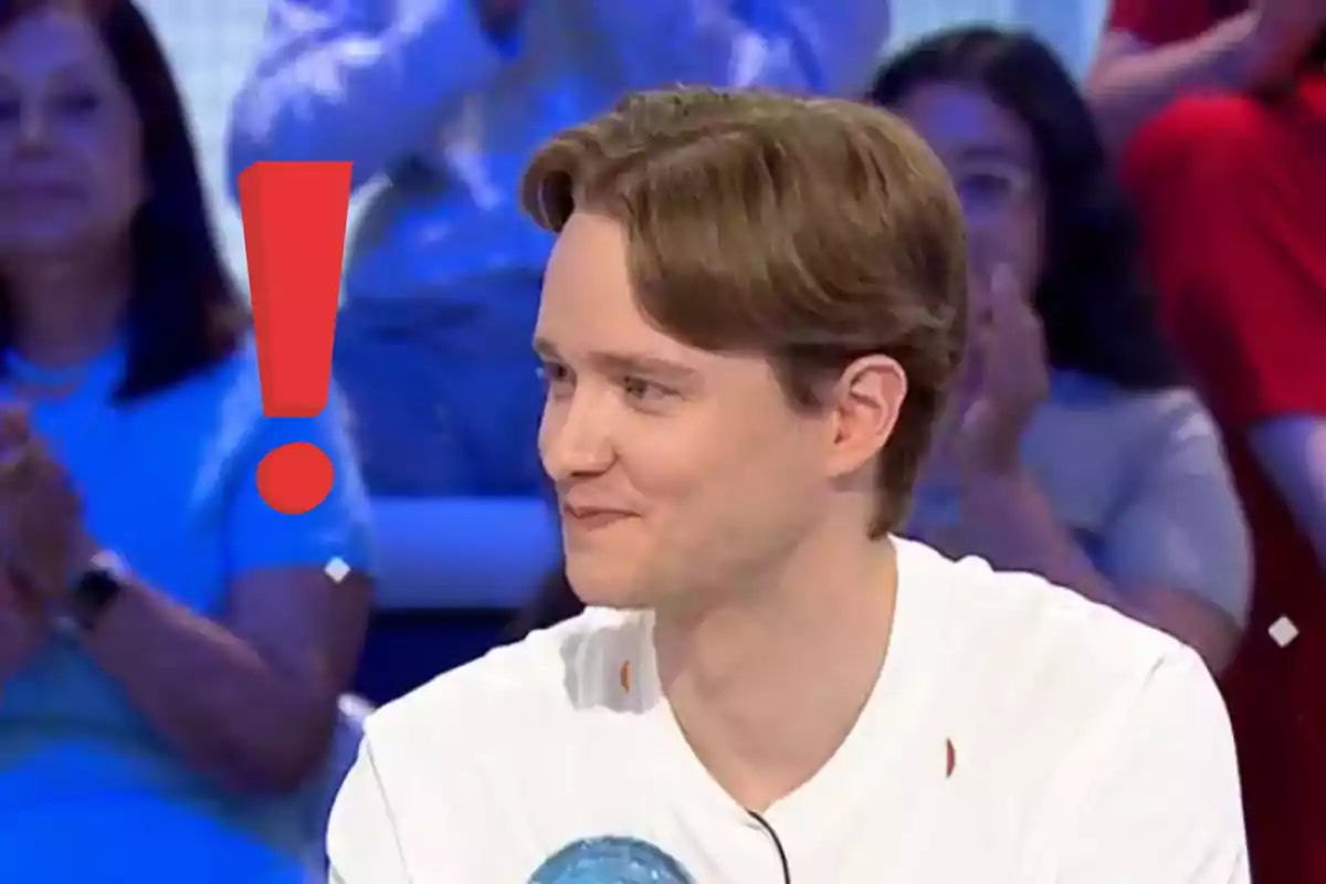 Young man with light brown hair smiles while sitting on a TV show with a large red exclamation mark next to him and blurred people in the background.