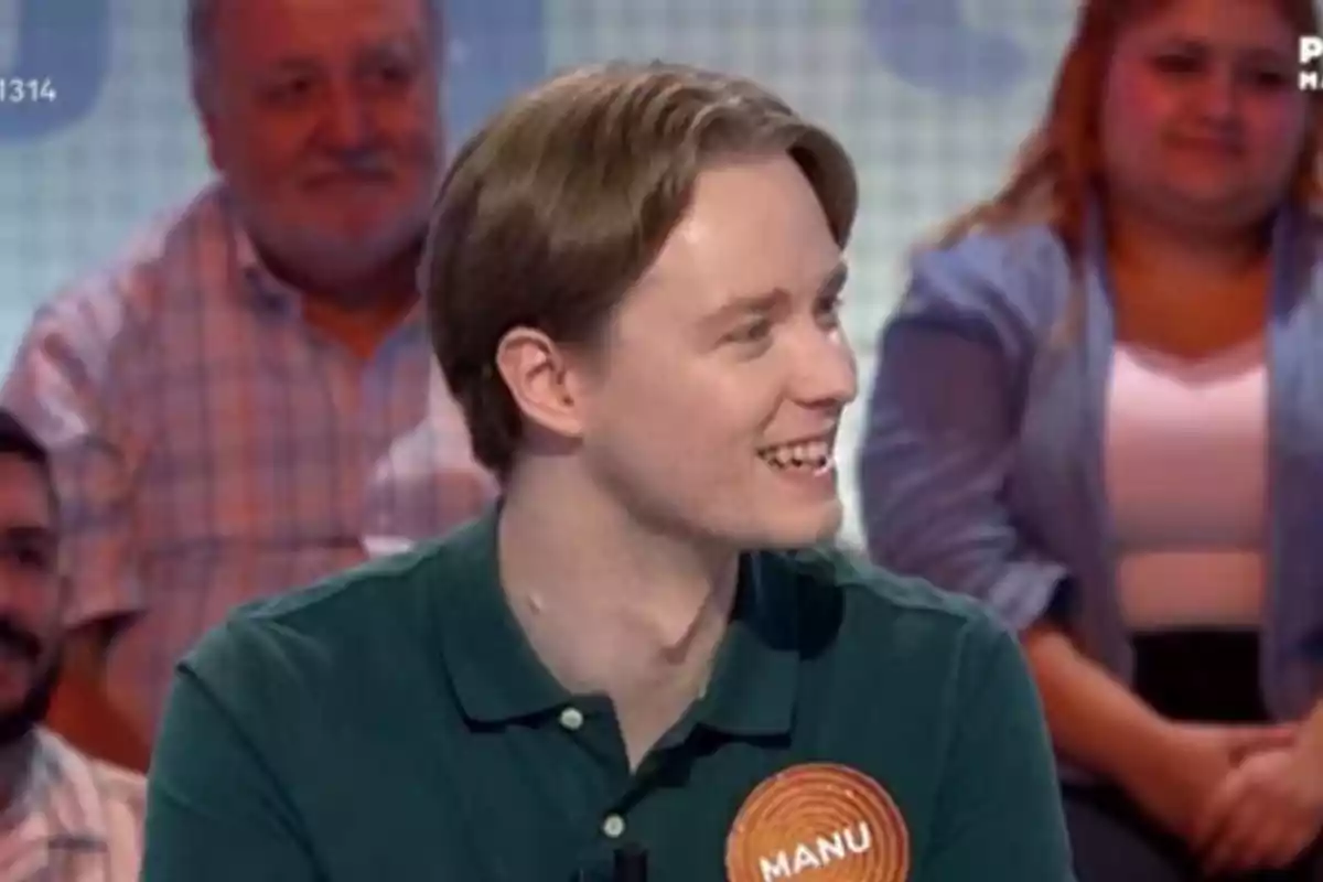 Un jove somrient amb una samarreta verda i una xapa que diu Manu participa en un programa de televisió amb públic al fons Un jove somrient amb una samarreta verda i una xapa que diu Manu participa en un programa de televisió amb públic al fons