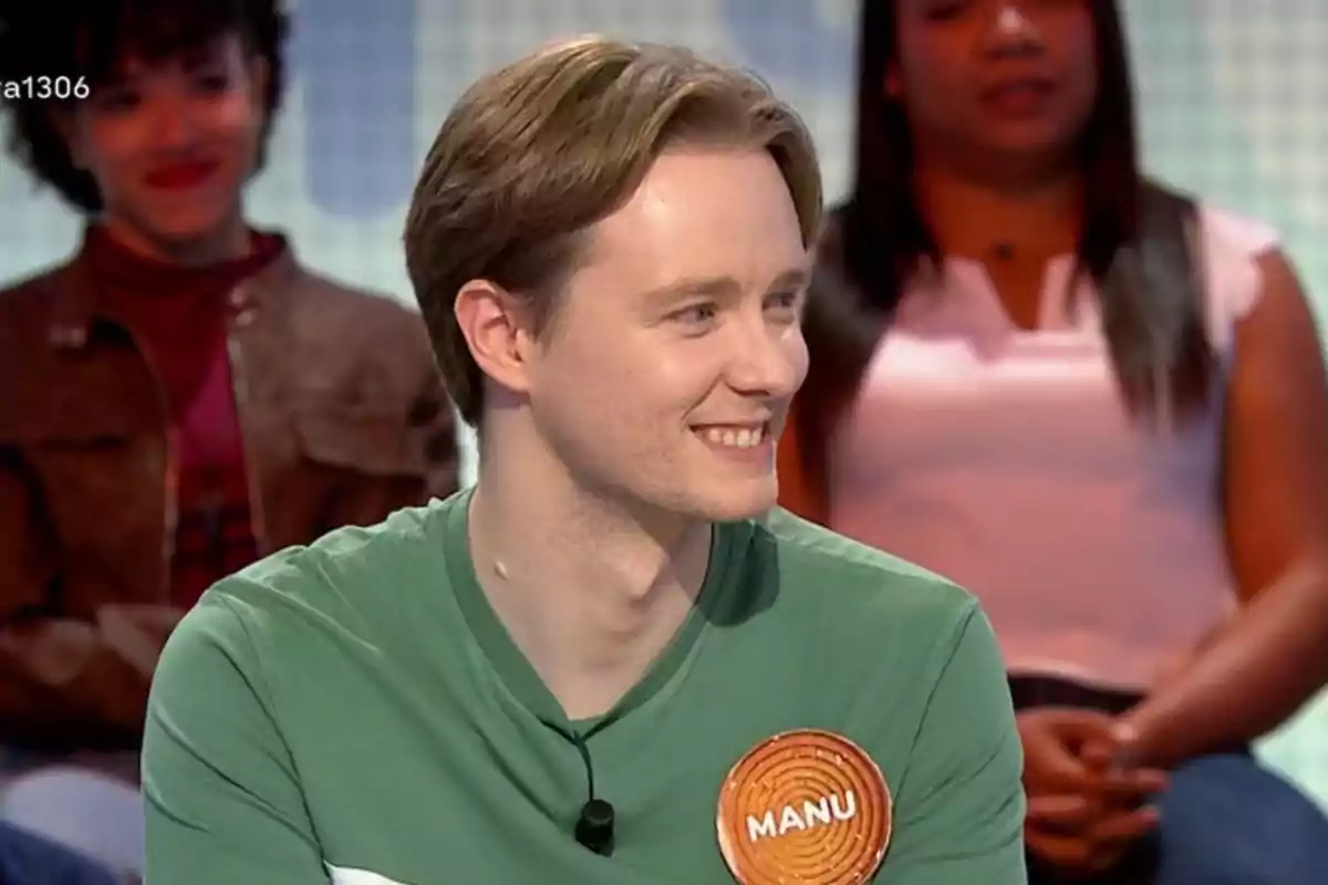 Manu sonriente con camiseta verde y un gafete que dice Manu, sentado en un set de televisión con personas al fondo. Manu sonriente con camiseta verde y un gafete que dice Manu, sentado en un set de televisión con personas al fondo.