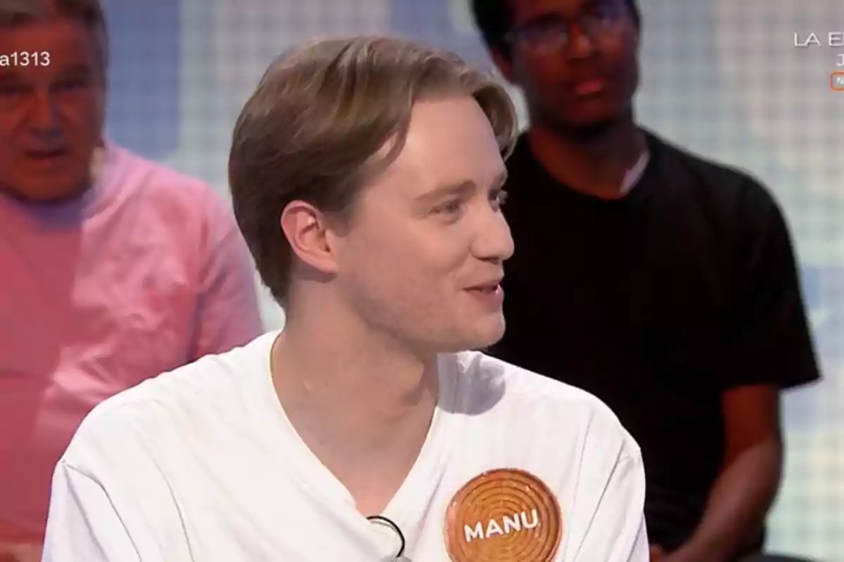 Manu con camiseta blanca y gafas colgadas conversando en un programa de televisión con una chapa que dice Manu y personas sentadas al fondo Manu con camiseta blanca y gafas colgadas conversando en un programa de televisión con una chapa que dice Manu y personas sentadas al fondo
