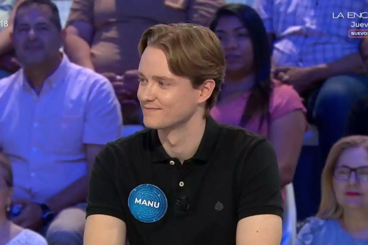 Un jove amb samarreta negra i una placa blava que diu Manu participa en un programa de concursos amb públic al fons. Un jove amb samarreta negra i una placa blava que diu Manu participa en un programa de concursos amb públic al fons.