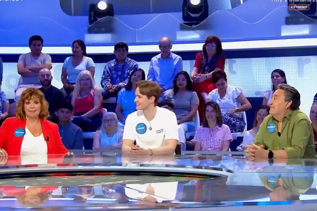 Three people sitting at a table on a game show with an audience in the background Three people sitting at a table on a game show with an audience in the background