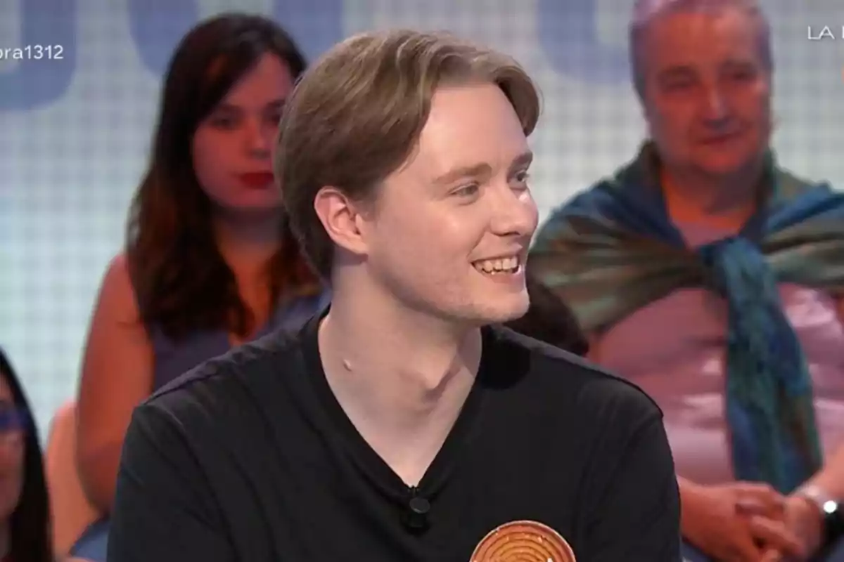 Un jove somrient amb samarreta negra participa en un programa de televisió amb públic al fons. Un jove somrient amb samarreta negra participa en un programa de televisió amb públic al fons.