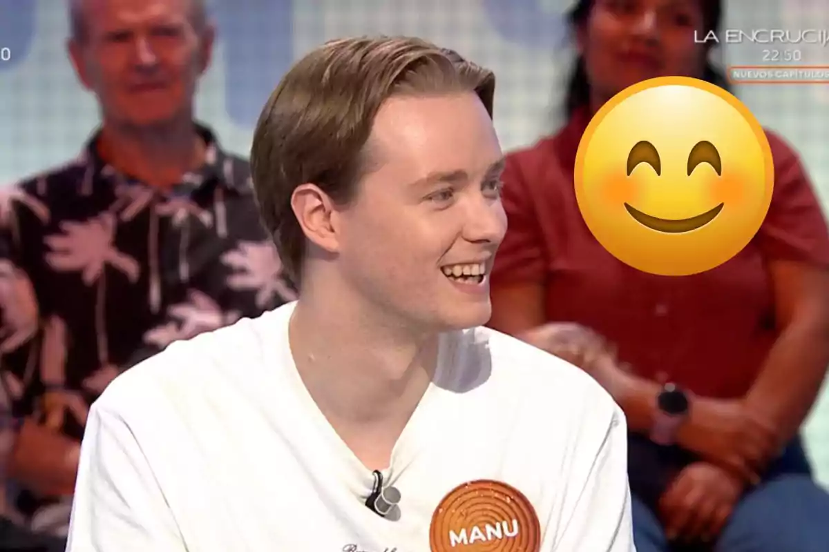 Un jove somrient amb una samarreta blanca i un pin que diu Manu participa en un programa de televisió mentre un emoji somrient cobreix parcialment una persona del públic al fons.