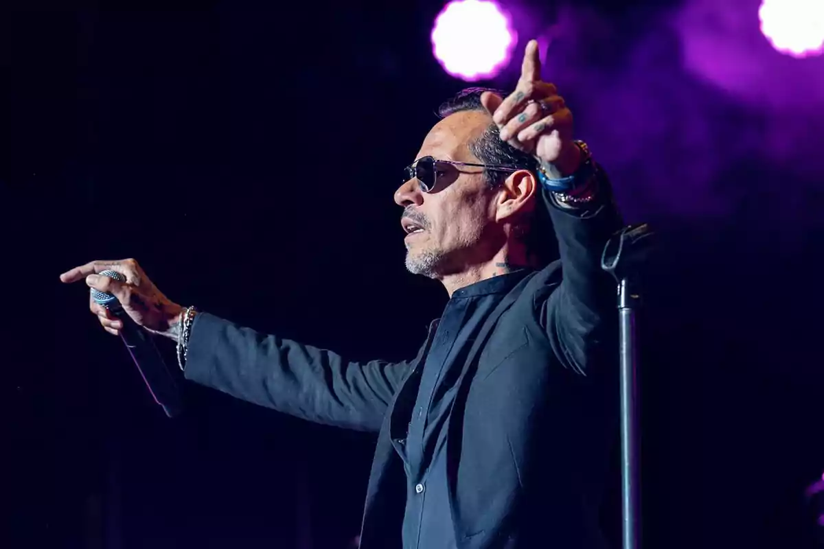 Marc Anthony con gafas de sol y micrófono en la mano cantando en un escenario iluminado con luces moradas Marc Anthony con gafas de sol y micrófono en la mano cantando en un escenario iluminado con luces moradas