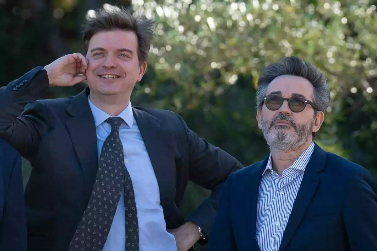 Dos hombres con traje posan al aire libre uno de ellos sonríe y el otro lleva gafas de sol Dos hombres con traje posan al aire libre uno de ellos sonríe y el otro lleva gafas de sol
