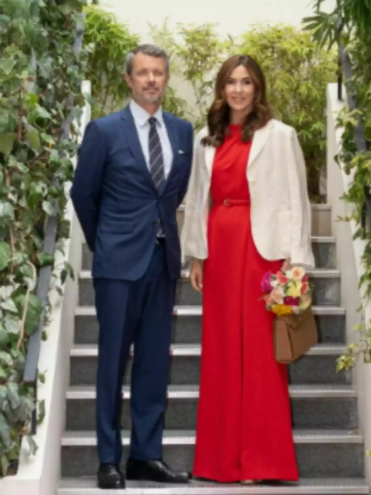 Couple consisting of Mary and Frederik of Denmark posing on a staircase surrounded by plants; the man is wearing a blue suit and the woman is dressed in a red outfit with a light-colored jacket and holding a bouquet of flowers. Couple consisting of Mary and Frederik of Denmark posing on a staircase surrounded by plants; the man is wearing a blue suit and the woman is dressed in a red outfit with a light-colored jacket and holding a bouquet of flowers.