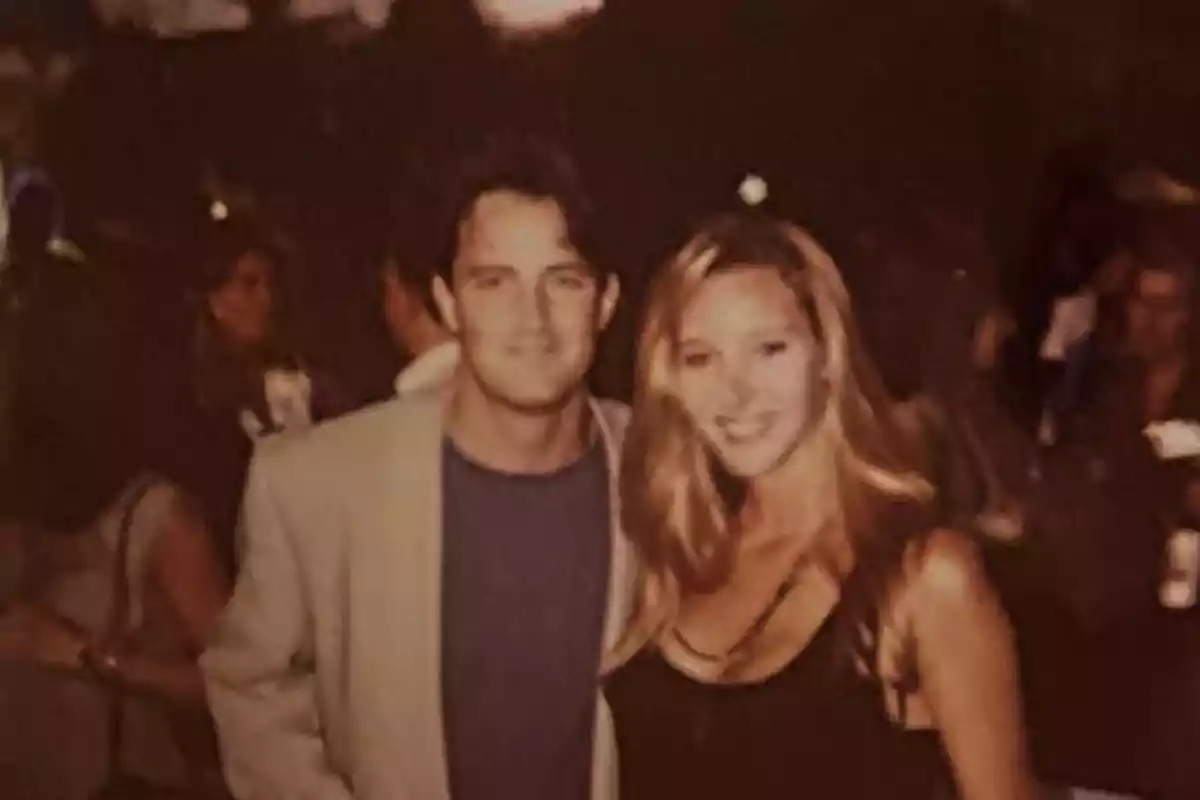 A couple smiles while posing for a photo at a social event with several people in the background. A couple smiles while posing for a photo at a social event with several people in the background.