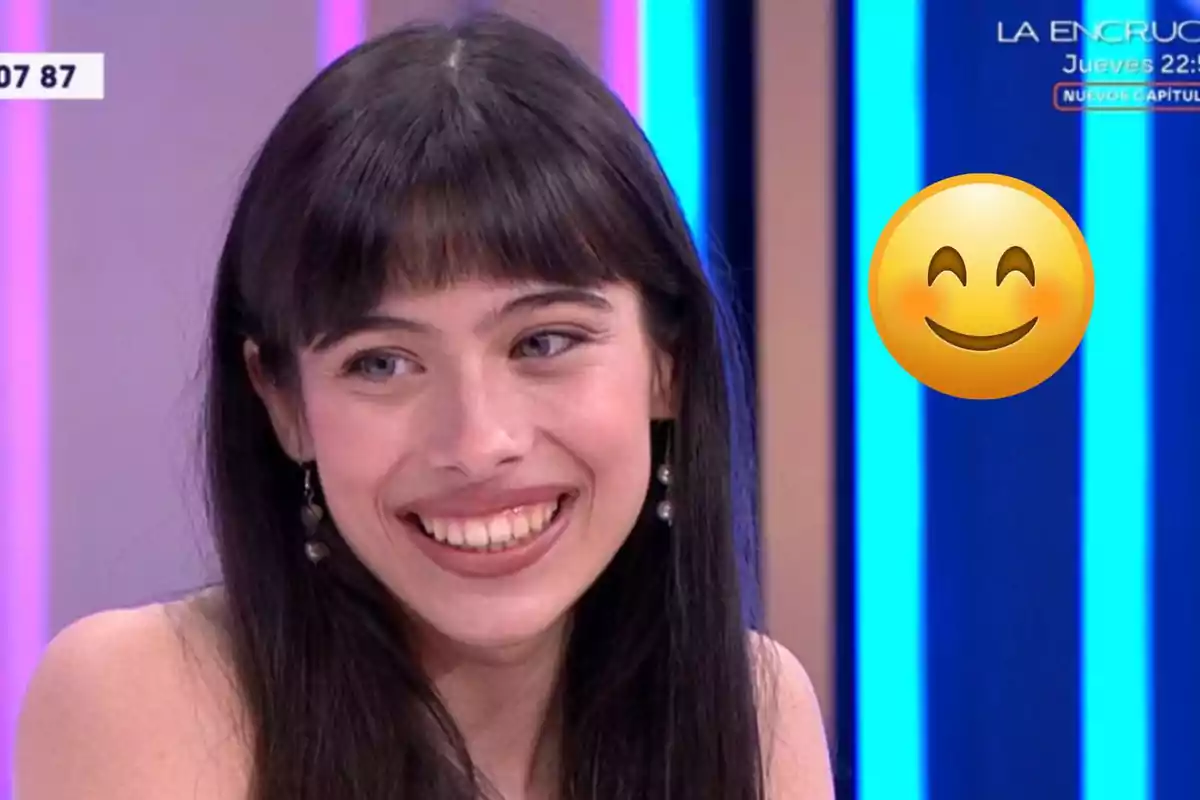 Melani de cabello largo y oscuro sonríe en un set de televisión con un emoji sonriente al lado