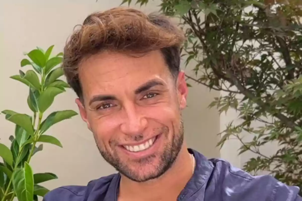 Montoya joven de cabello castaño claro y barba corta sonriendo frente a unas plantas Montoya joven de cabello castaño claro y barba corta sonriendo frente a unas plantas