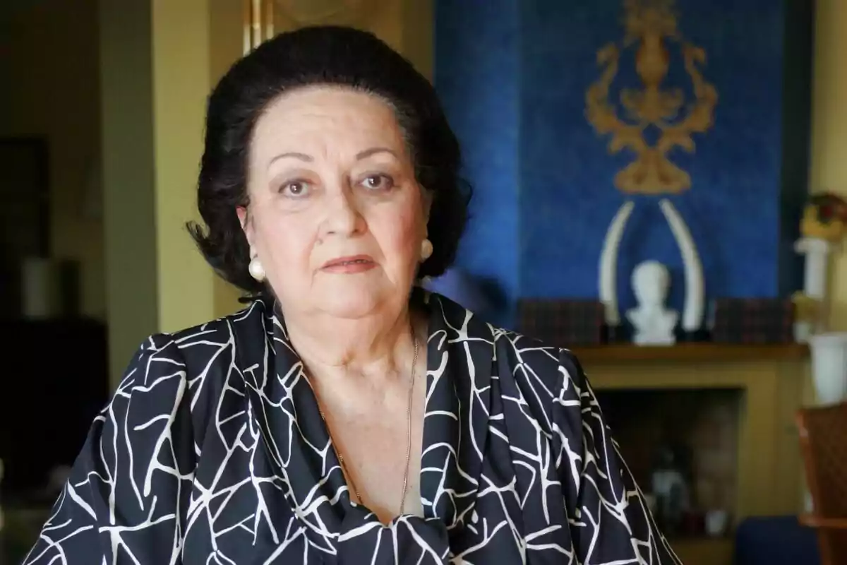 Dona gran amb cabell fosc i pentinat voluminós, amb arracades de perla i brusa estampada en blanc i negre, asseguda en una sala elegant amb una xemeneia i decoració blava al fons. Dona gran amb cabell fosc i pentinat voluminós, amb arracades de perla i brusa estampada en blanc i negre, asseguda en una sala elegant amb una xemeneia i decoració blava al fons.