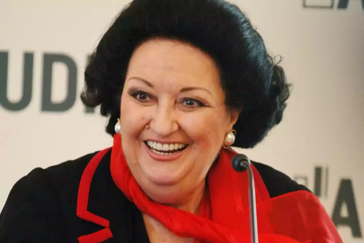 Dona de cabell fosc i curt somrient amb una bufanda vermella i arracades de perla davant d’un micròfon Dona de cabell fosc i curt somrient amb una bufanda vermella i arracades de perla davant d’un micròfon