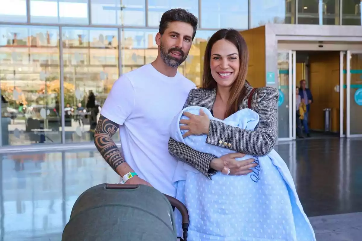 Una pareja sonriente está de pie frente a un edificio de cristal, la mujer sostiene a un bebé envuelto en una manta azul y el hombre lleva una camiseta blanca y tiene un cochecito de bebé. Una pareja sonriente está de pie frente a un edificio de cristal, la mujer sostiene a un bebé envuelto en una manta azul y el hombre lleva una camiseta blanca y tiene un cochecito de bebé.