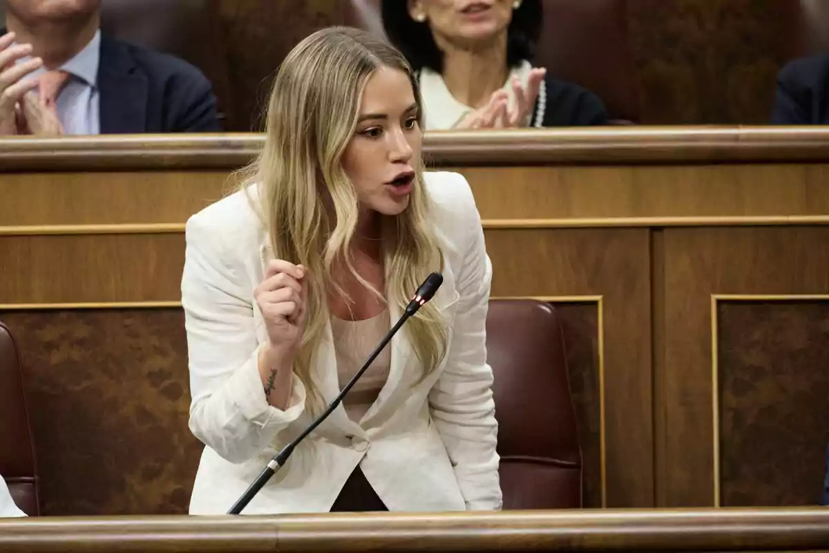 Mujer joven de cabello rubio y suelto hablando con énfasis frente a un micrófono en un recinto parlamentario Mujer joven de cabello rubio y suelto hablando con énfasis frente a un micrófono en un recinto parlamentario