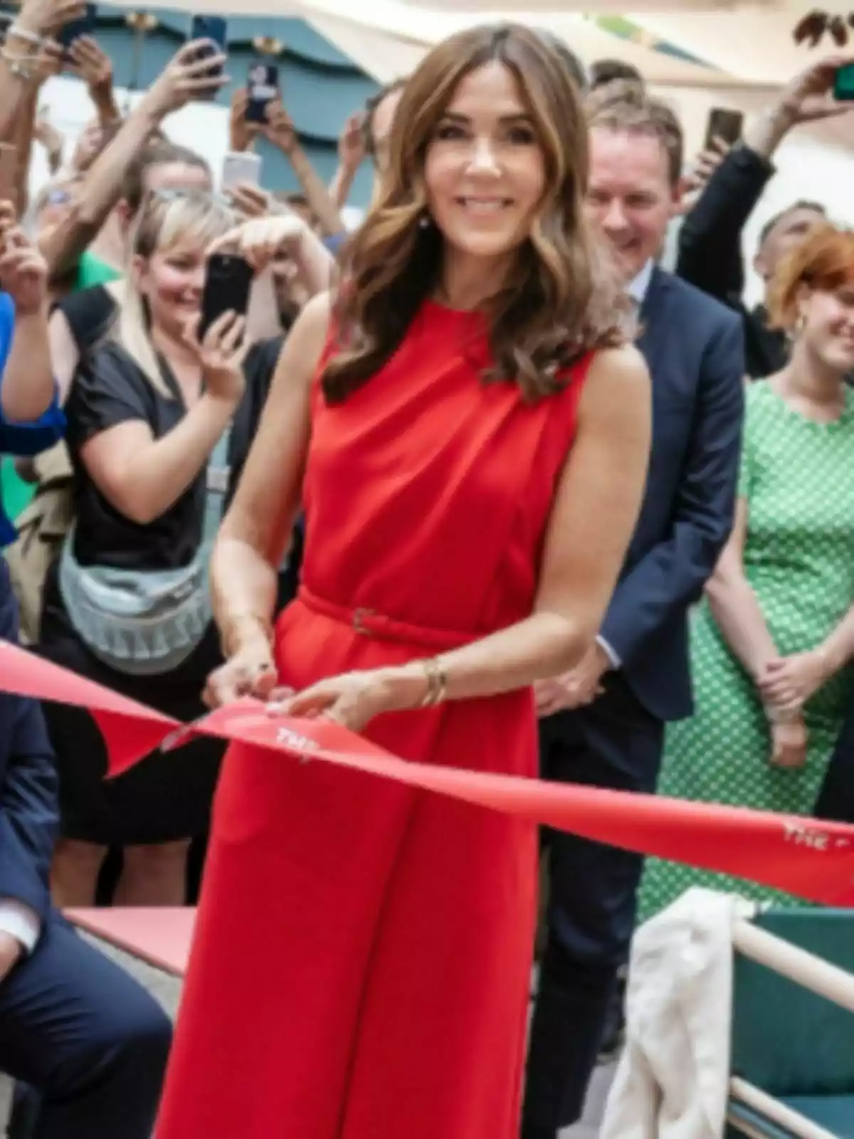 Mary of Denmark in a red dress cutting a ribbon at an opening ceremony while several people watch and take photos. Mary of Denmark in a red dress cutting a ribbon at an opening ceremony while several people watch and take photos.