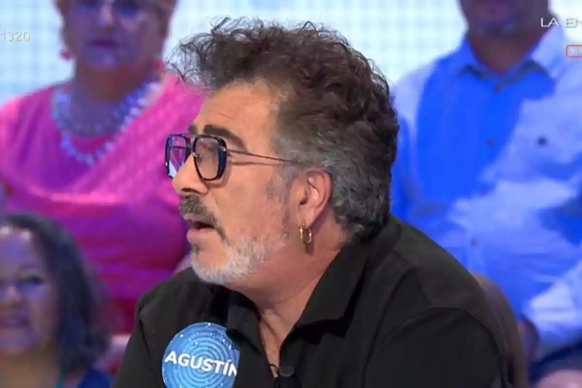Home de cabells arrissats i ulleres negres participant en un programa de televisió amb una etiqueta blava que diu Agustín i persones assegudes al fons