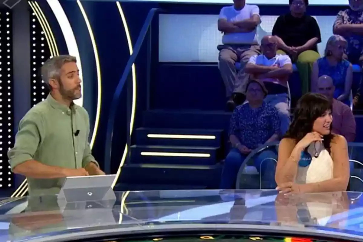 Roberto Leal de pie junto a una mesa transparente y una concursante sentada en un plató de televisión con público al fondo Roberto Leal de pie junto a una mesa transparente y una concursante sentada en un plató de televisión con público al fondo