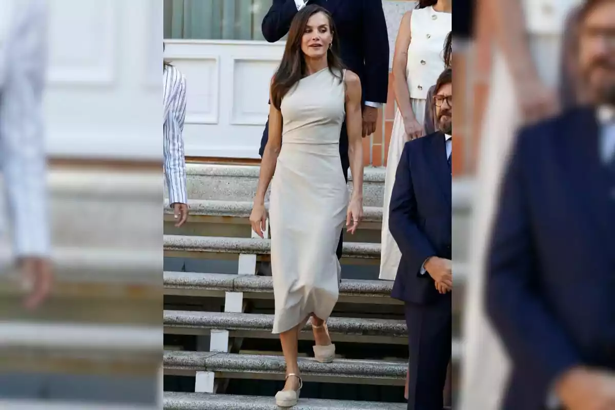Mujer con vestido beige sin mangas bajando unas escaleras rodeada de otras personas elegantemente vestidas Mujer con vestido beige sin mangas bajando unas escaleras rodeada de otras personas elegantemente vestidas