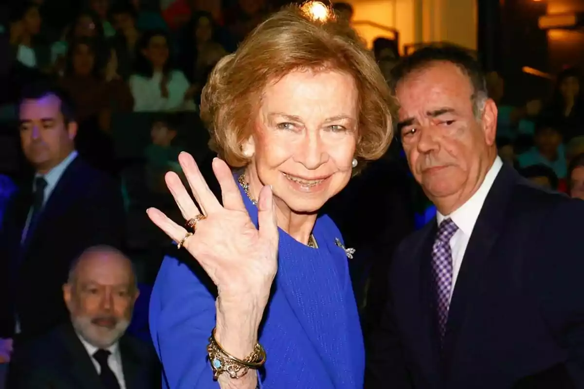 Dona gran amb vestit blau saludant amb la mà i somrient mentre diverses persones l’envolten en un esdeveniment formal Dona gran amb vestit blau saludant amb la mà i somrient mentre diverses persones l’envolten en un esdeveniment formal