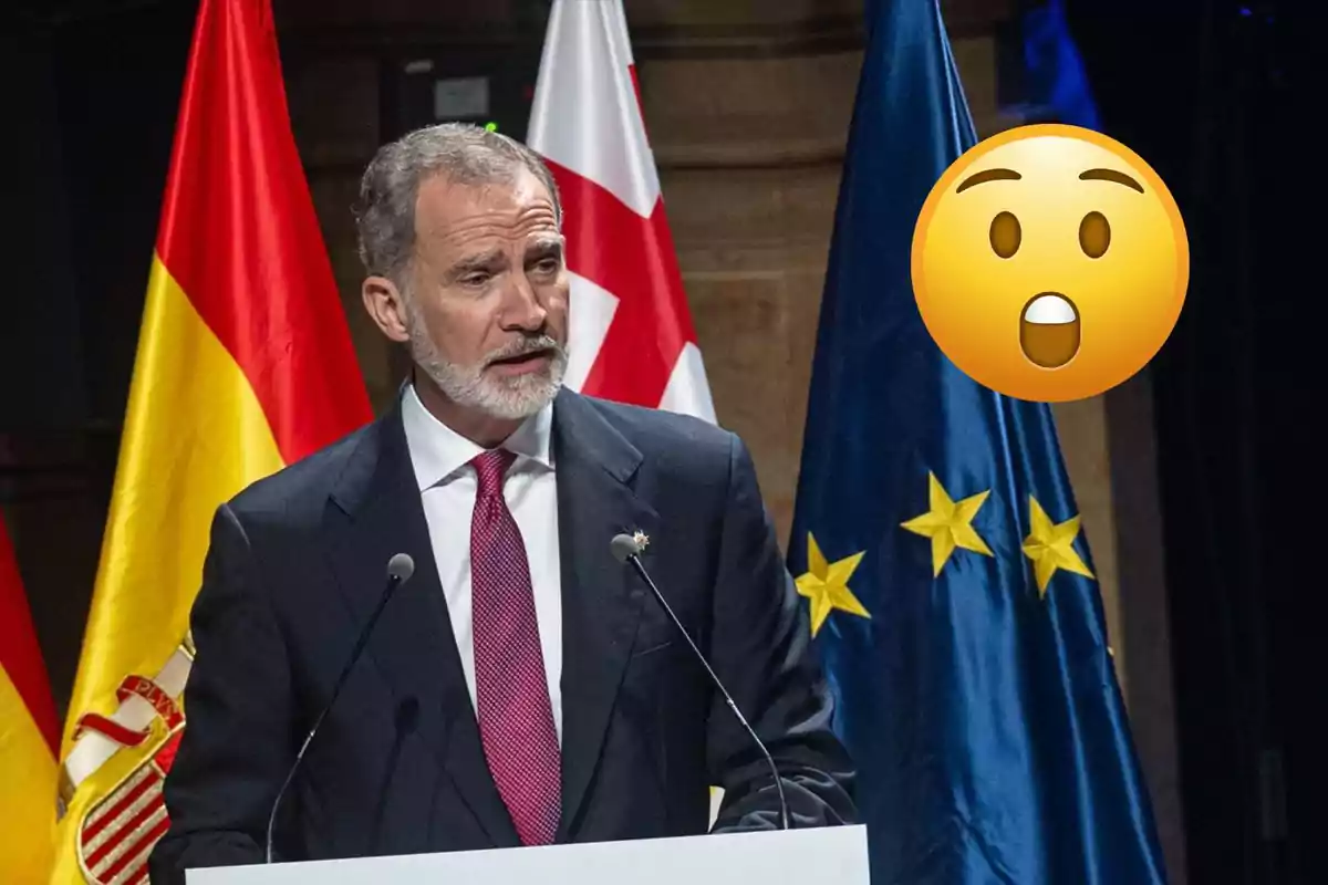 Felipe VI de traje hablando en un podio con banderas de España y la Unión Europea de fondo y un emoji de sorpresa grande a su lado