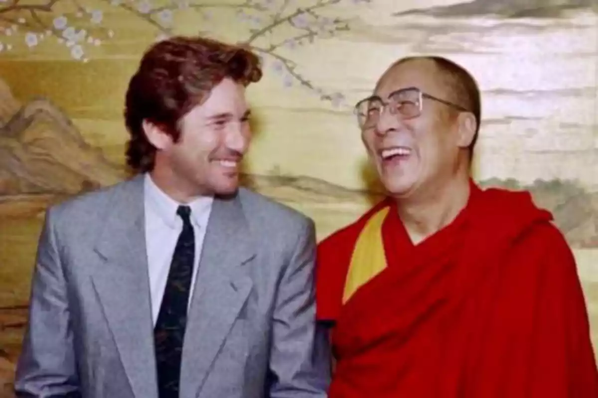 Richard Gere y el Dalái Lama sonríen y se miran con alegría, uno lleva un traje gris y el otro viste una túnica roja con detalles amarillos, posando frente a un fondo decorativo. Richard Gere y el Dalái Lama sonríen y se miran con alegría, uno lleva un traje gris y el otro viste una túnica roja con detalles amarillos, posando frente a un fondo decorativo.