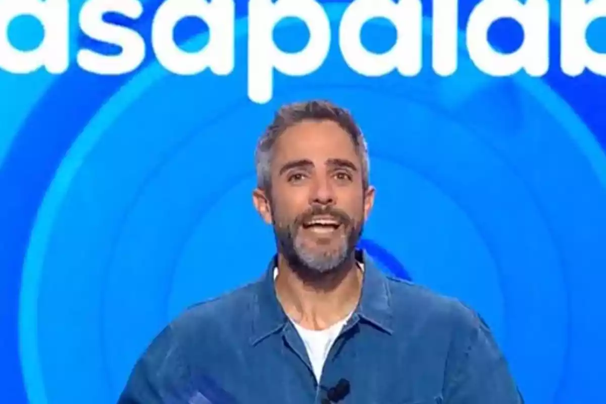 Roberto Leal con barba y cabello corto canoso sonríe frente a un fondo azul con letras blancas grandes