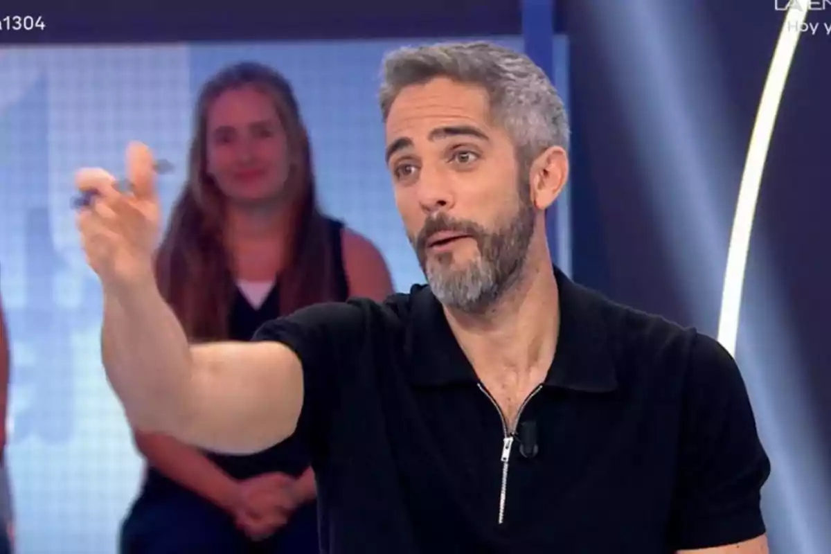 Gray-haired, bearded man dressed in a black t-jersey speaking and gesturing on a television set with a blurred woman in the background Gray-haired, bearded man dressed in a black t-jersey speaking and gesturing on a television set with a blurred woman in the background