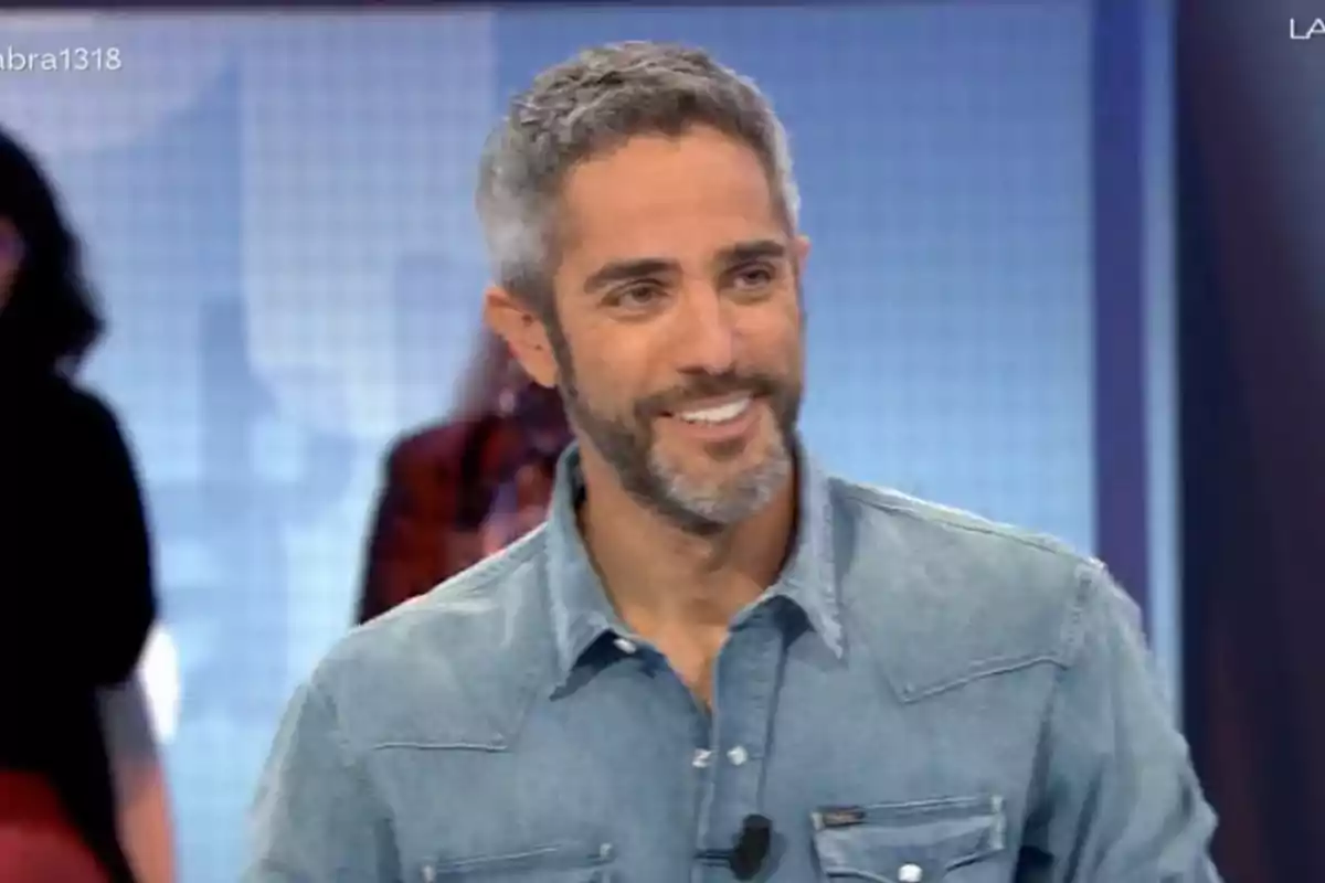 Home de cabell curt i barba canosa somrient mentre porta una camisa texana en un estudi de televisió Home de cabell curt i barba canosa somrient mentre porta una camisa texana en un estudi de televisió