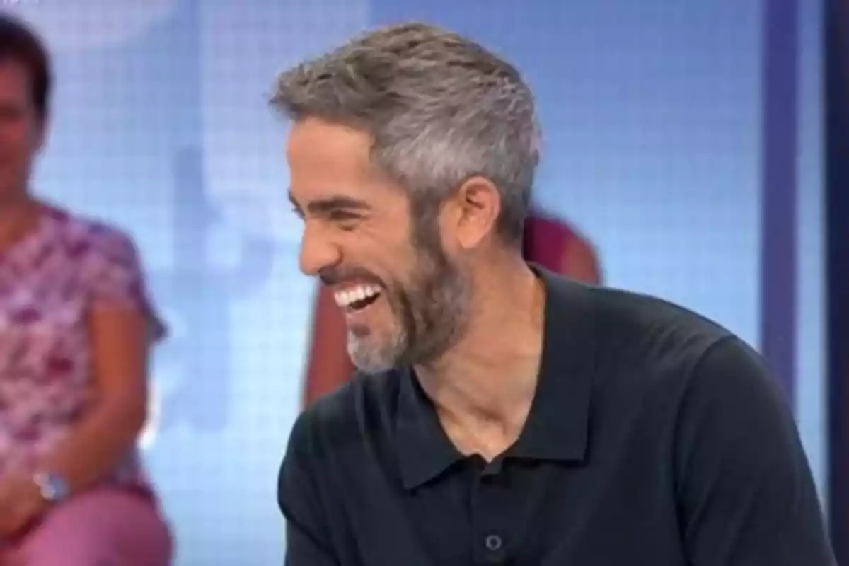 Home de cabells canosos i barba rient en un estudi de televisió amb persones desenfocades al fons Home de cabells canosos i barba rient en un estudi de televisió amb persones desenfocades al fons